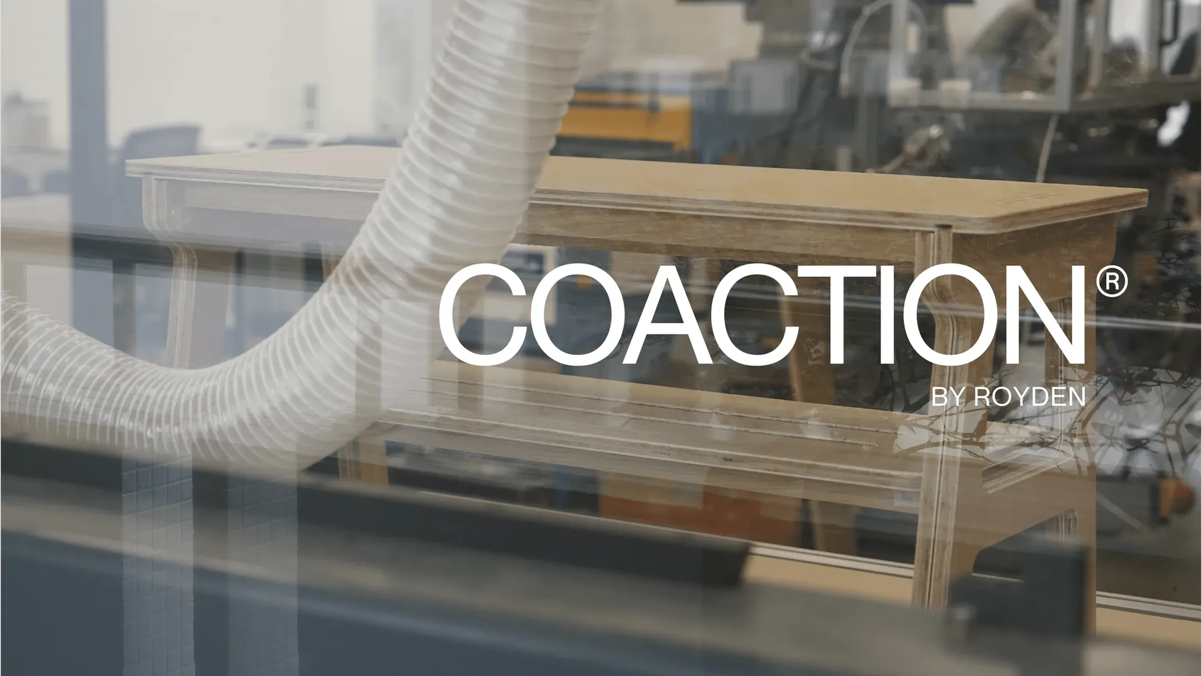 The white COACTION® BY ROYDEN wordmark over a hazy photo of the plywood shoe bench in a school workshop, seen through glass