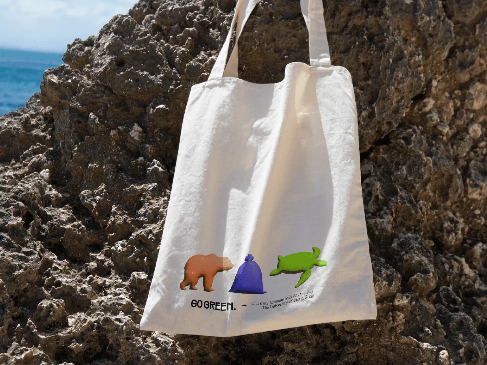 A canvas Go Green tote printed with the bear, bag and turtle, hanging against coastal rocks