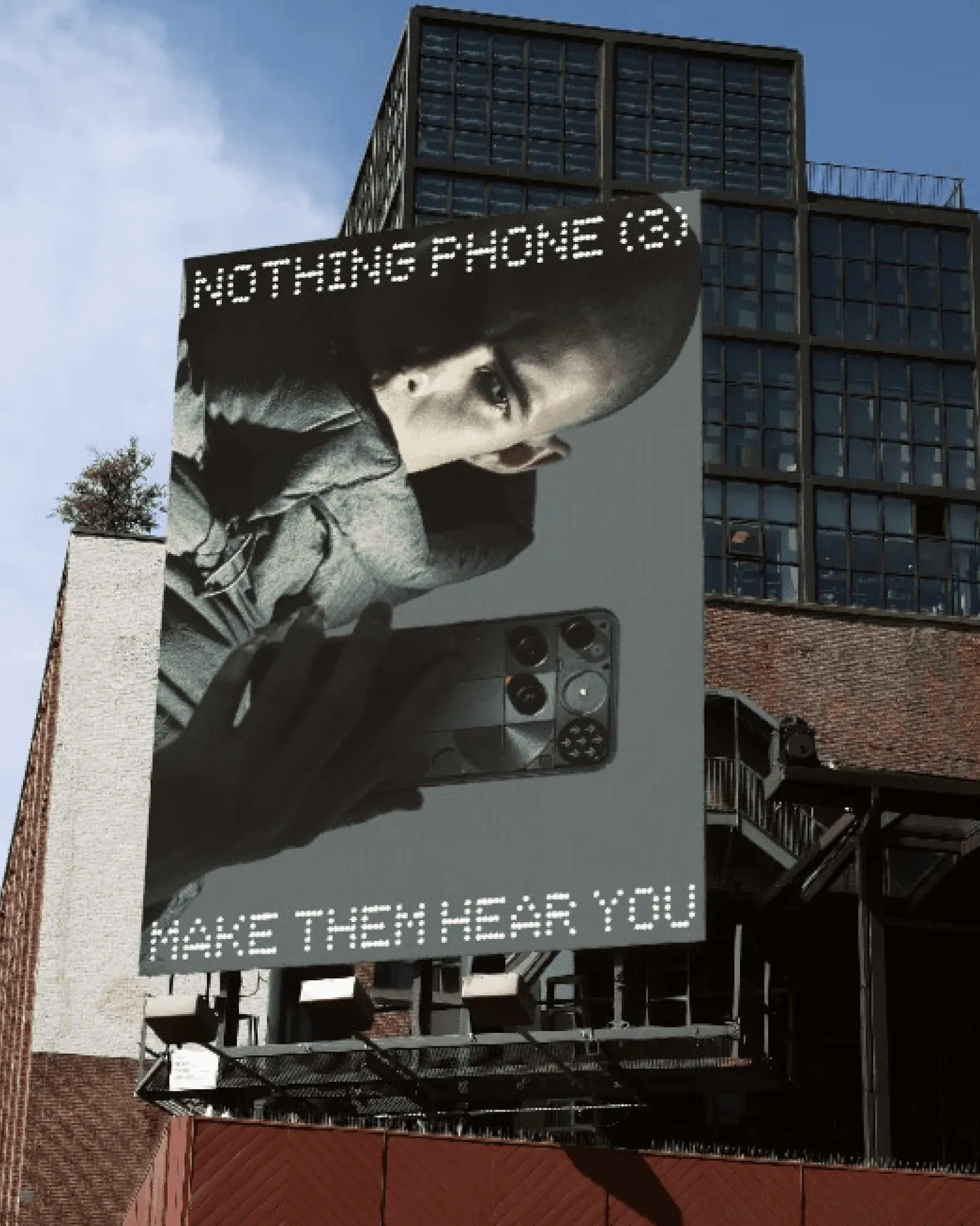 A rooftop billboard against blue sky: a man holding a phone, NOTHING PHONE (3) and MAKE THEM HEAR YOU in dot type