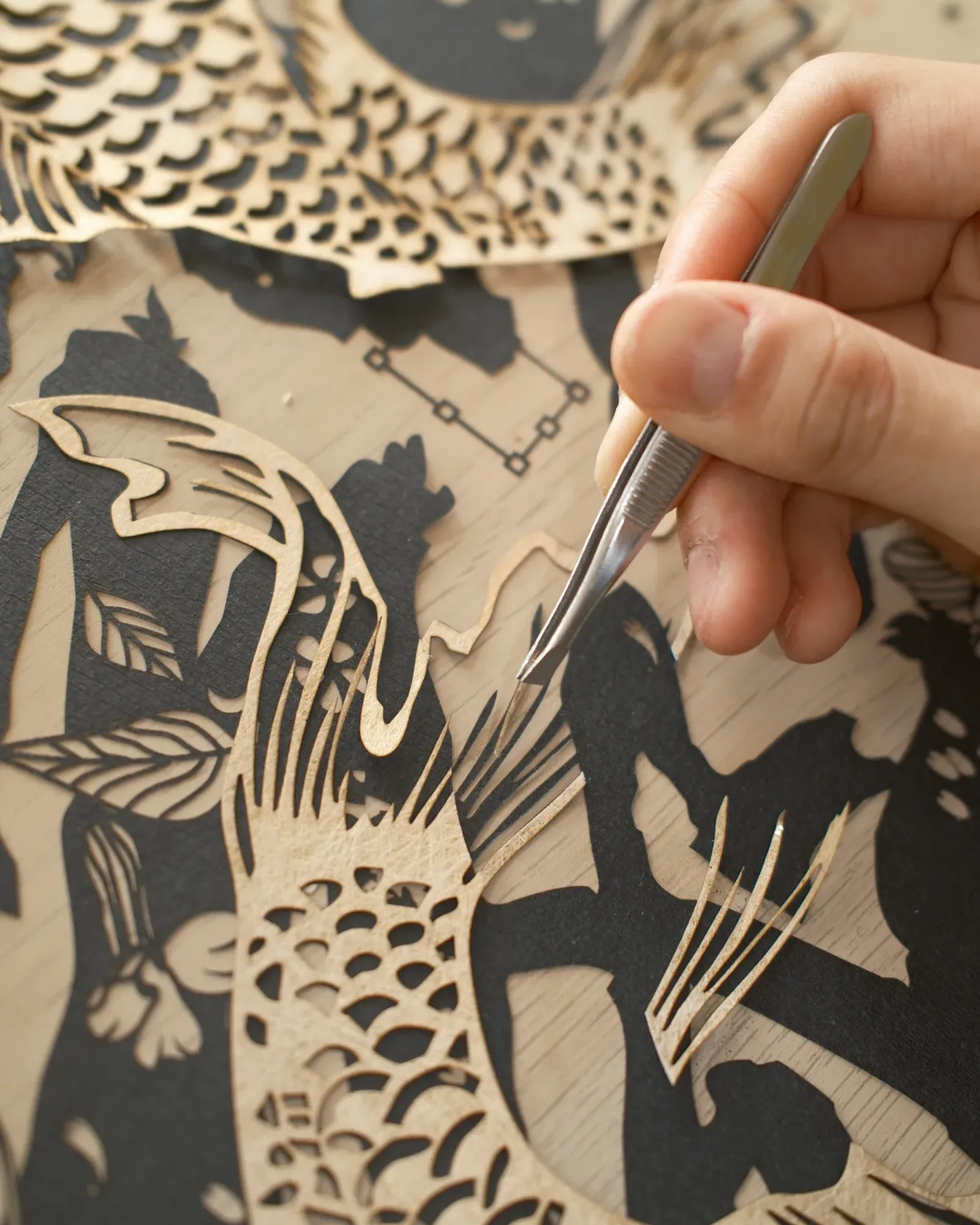 A hand placing gold papercut koi scales with tweezers on black papercut