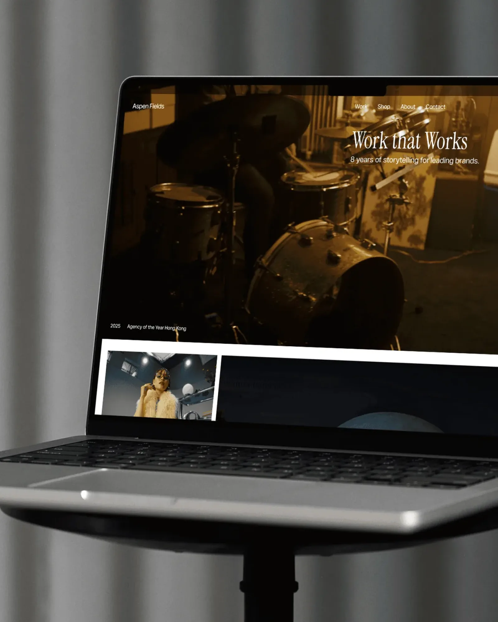 A MacBook on a stool in front of a grey curtain, showing the Fields homepage: Work that Works over a warm film of a drummer