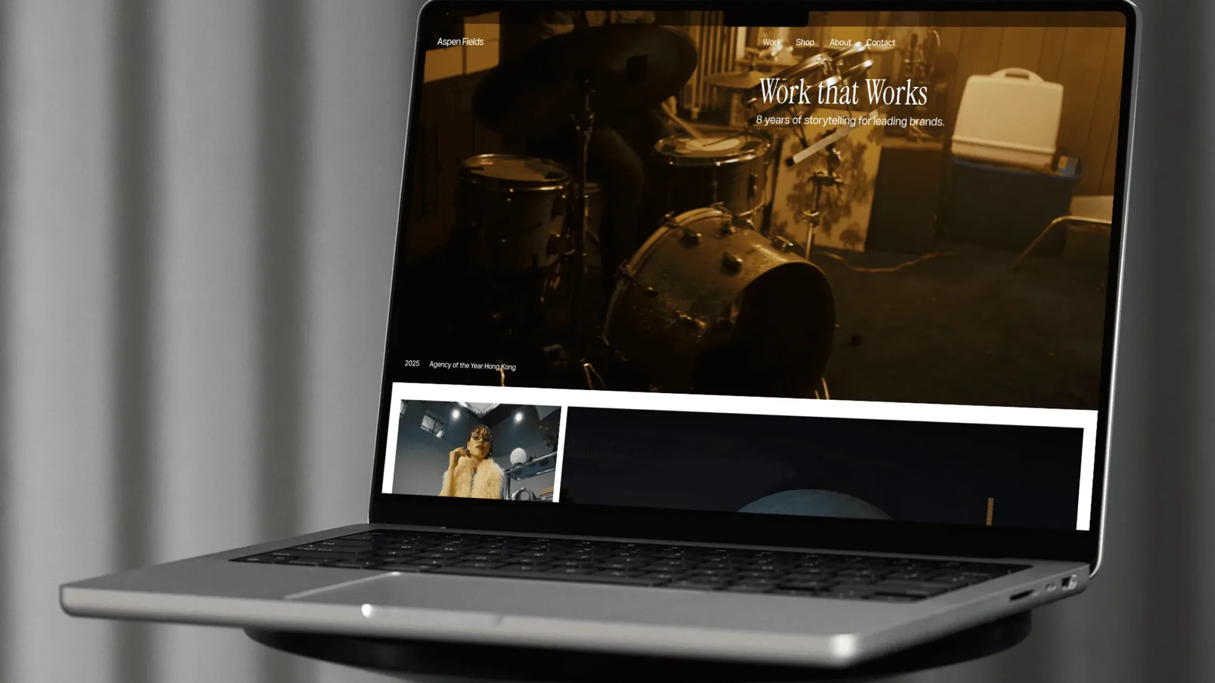 A MacBook on a stool in front of a grey curtain, showing the Fields homepage: Work that Works over a warm film of a drummer