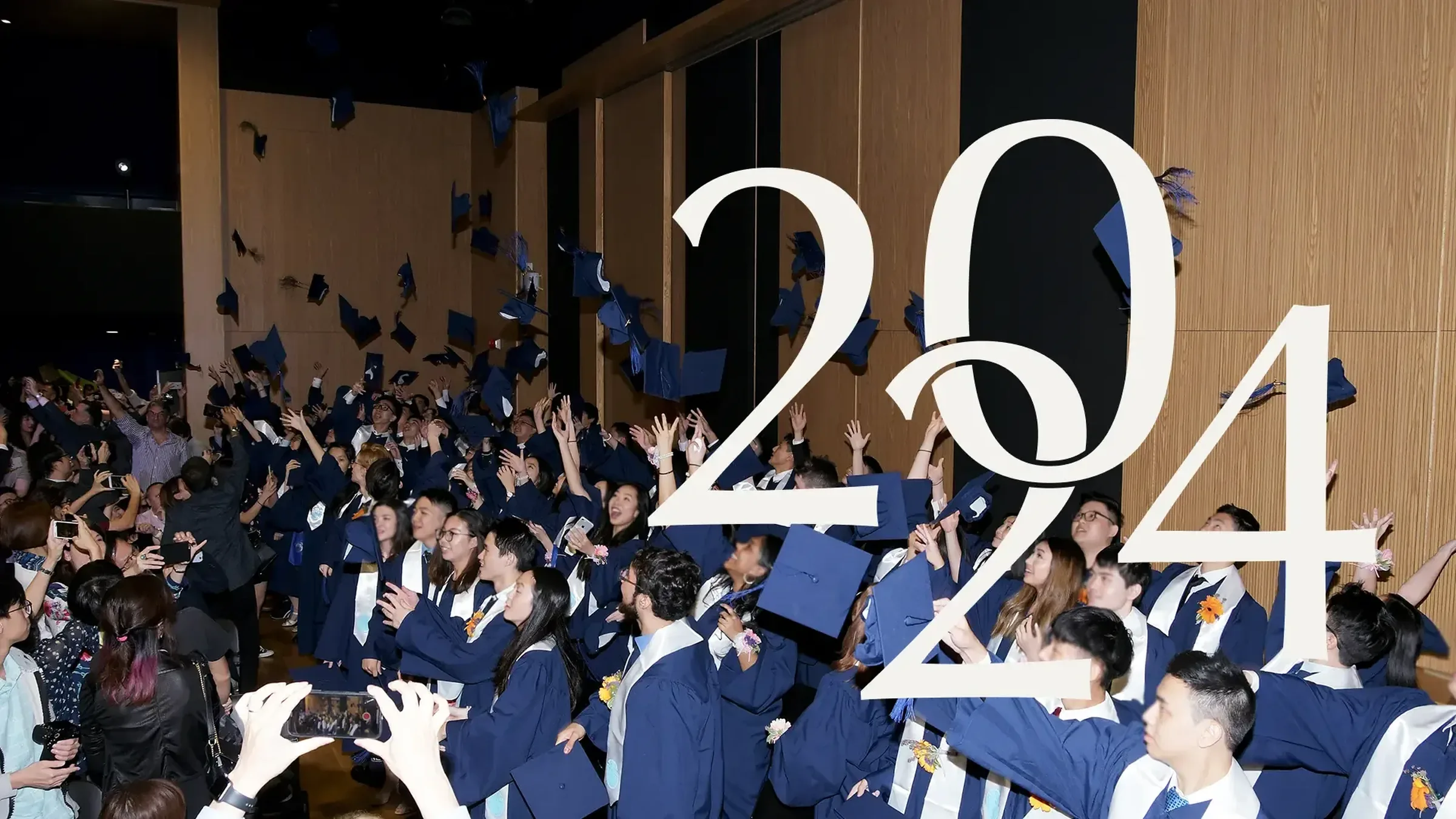 Graduates in navy gowns throwing their caps in the air, with the cream interlocking 2024 monogram over them