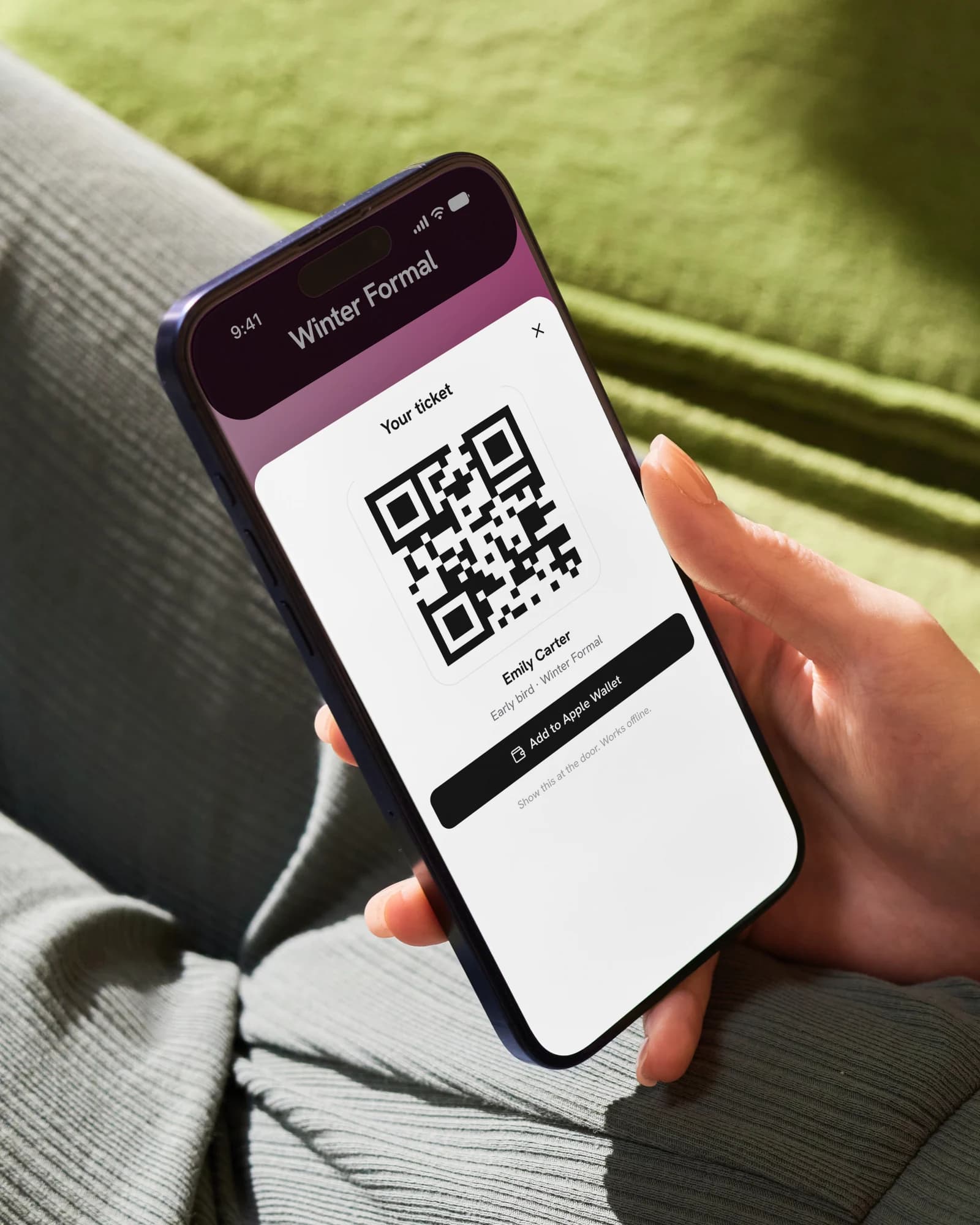 An iPhone held on a lap over a green throw: a guest's ticket, a QR code and Add to Apple Wallet