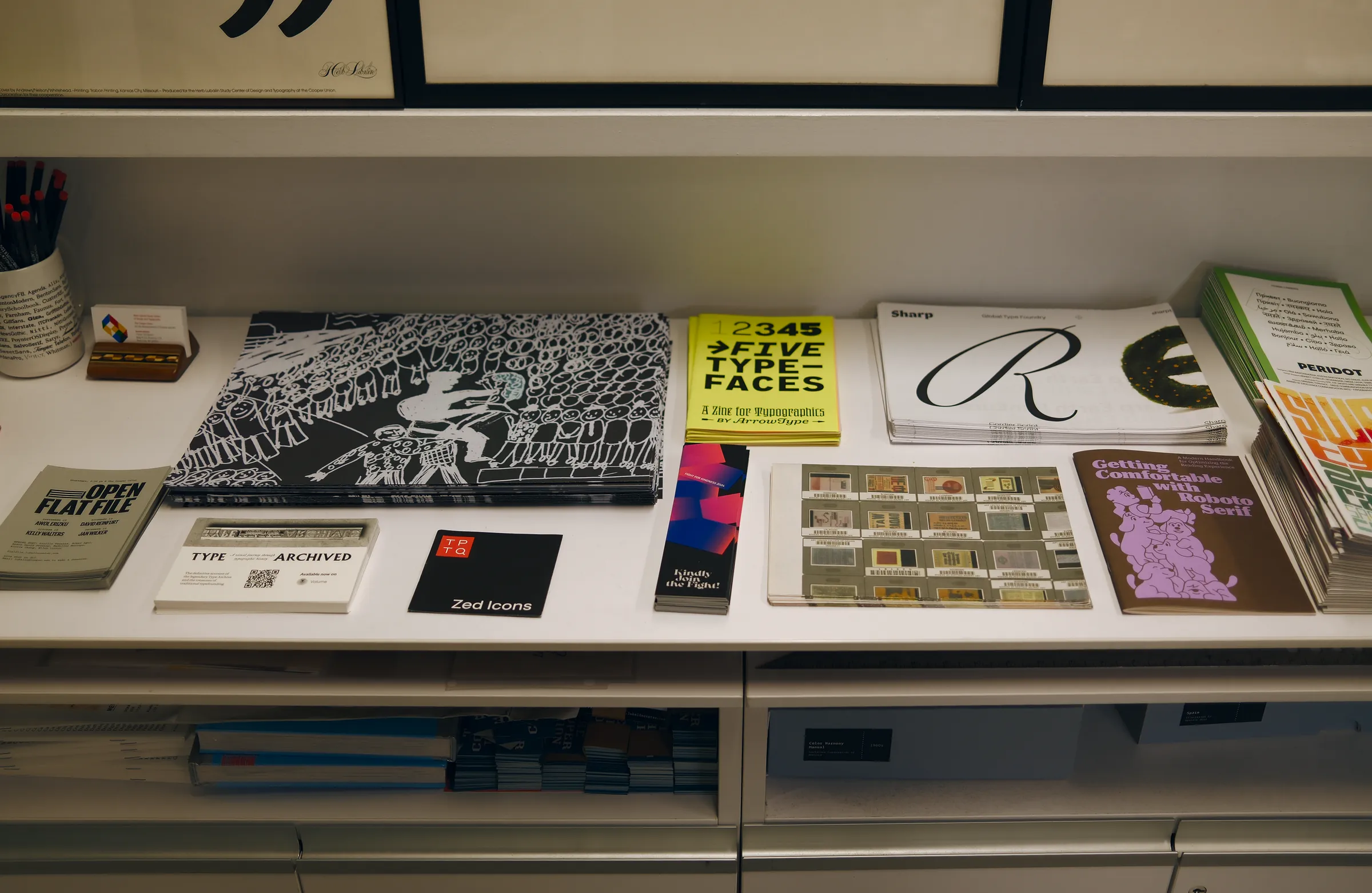 Royden's desk: zines, type specimens and printed matter laid out on a white shelf under framed posters.